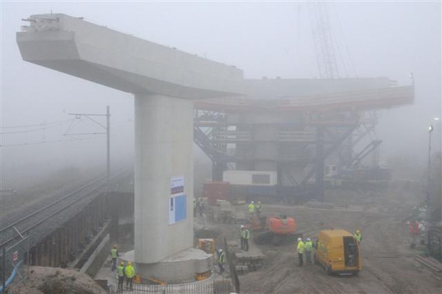 Foto Ronald Bakker
Medewerkers van aannemersbedrijf Heijmans draaien dinsdagmorgen in de mist een dwarsligger voor de toekomstige Westrandweg op zijn plek. Een deel van de dwarsligger hangt over een goederenspoorbaan in Westpoort.