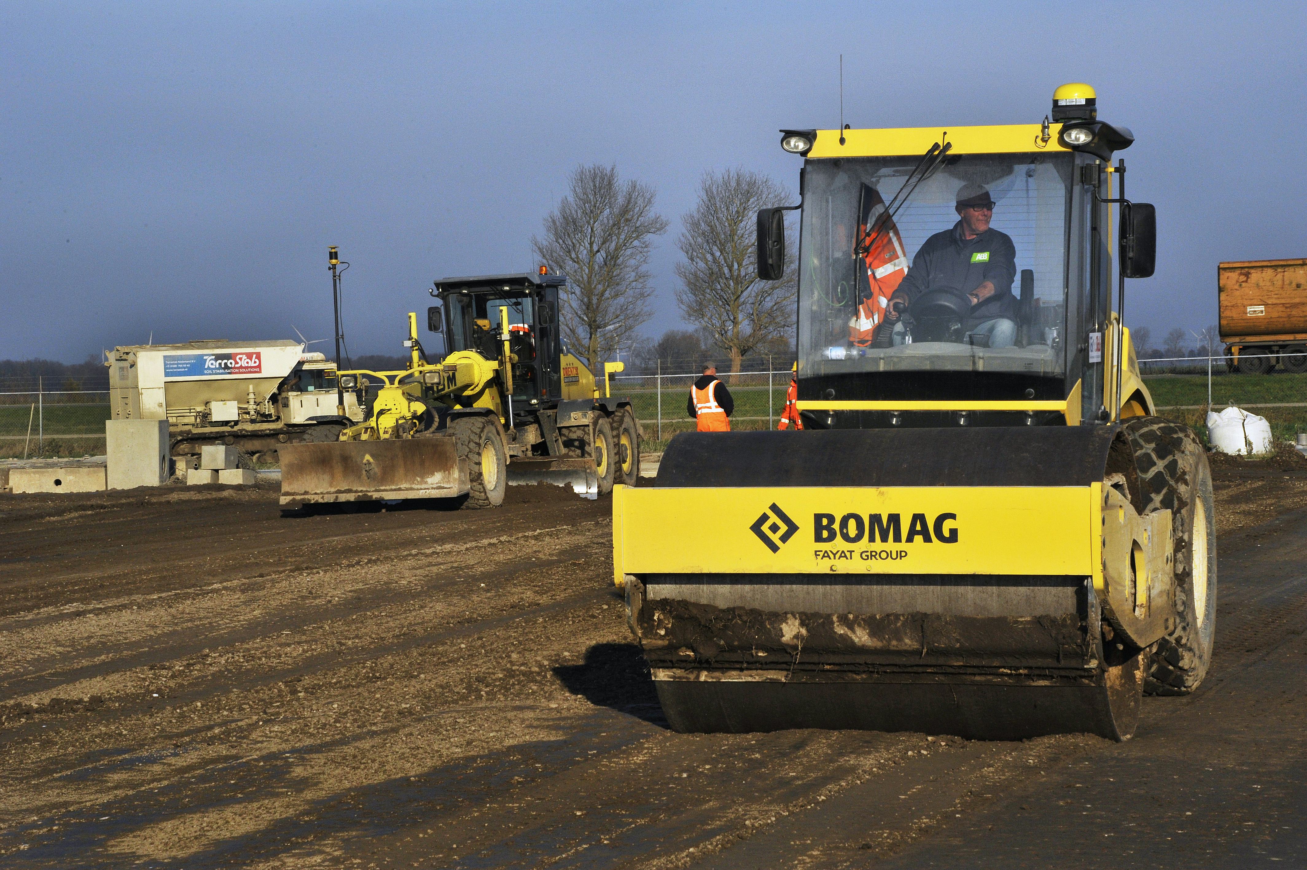 Een slimme wals en een grader egaliseren de kleibodem voor de nieuwe startbaan van Lelystad Airport. Met bindmiddelen is de bovenste laag natuurlijke klei zo hard gemaakt, dat een Boeing 737 er probleemloos op kan landen. Met een omvang van 300.000 m2 is Lelystad de eerste luchthaven in Nederland, waarbij bodemstabilisatie met succes op grote schaal is toegepast. Op de achtergrond de strooiwagen voor de bindmiddelen.
