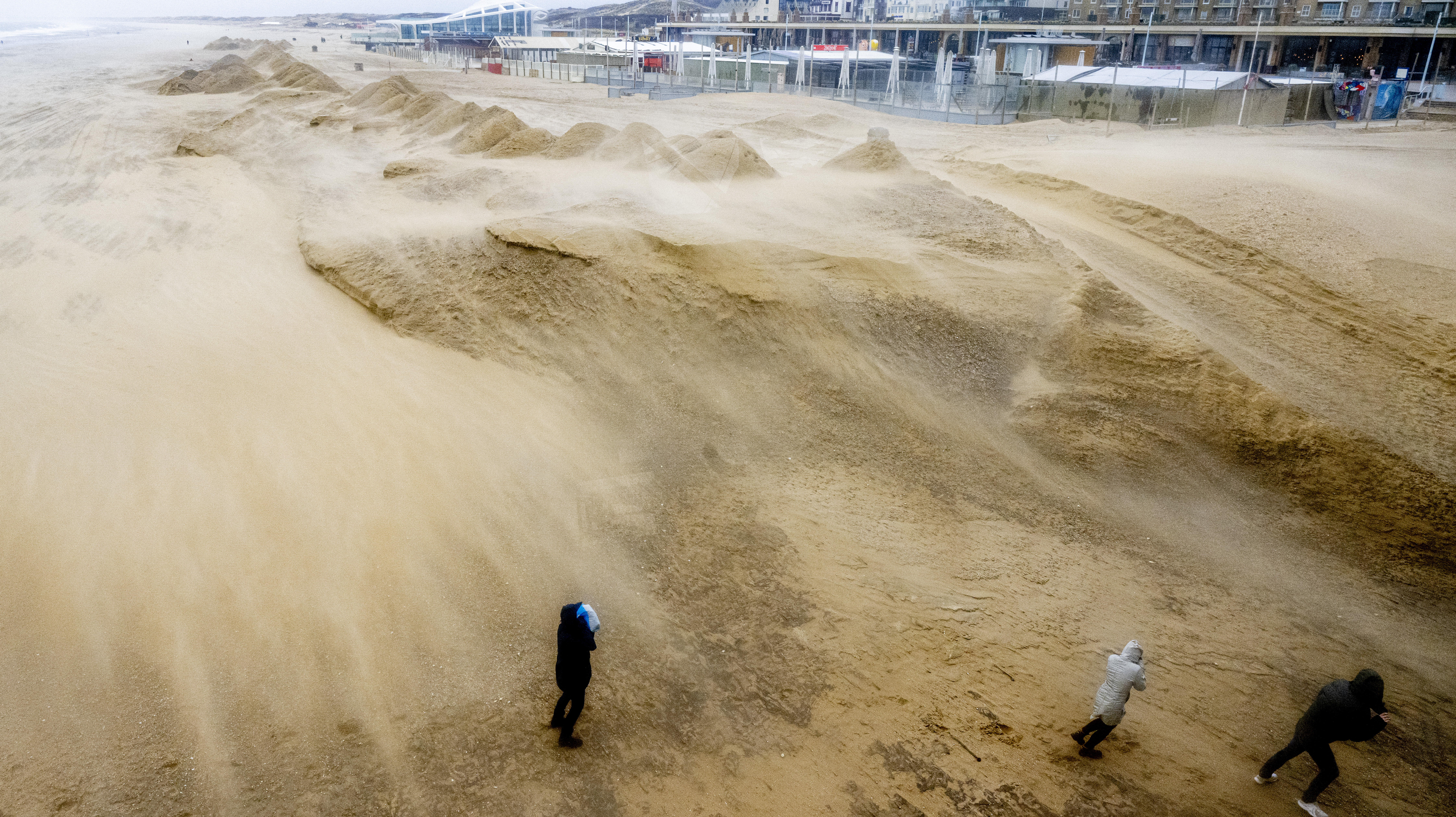 Mensen lopen door de harde wind langs een zandheuvel die strandtenten moet beschermen op het strand van Scheveningen.  ANP/Hollandse/Hoogte Robin Utrecht